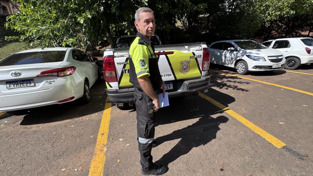 A imagem mostra um homem vestido com uniforme de alta visibilidade, predominantemente amarelo neon com preto, usando calça com faixas refletivas. Ele está segurando um papel branco e um rádio comunicador, olhando diretamente para a câmera. O cenário é um estacionamento ao ar livre, com várias árvores verdes ao fundo, indicando um ambiente aberto e ensolarado. Atrás do homem, há uma caminhonete Toyota Hilux branca, com detalhes em amarelo e branco na traseira formando um padrão em zigue-zague. Na lateral da caminhonete, está escrito "TRÂNSITO PIRACICABA", sugerindo que o veículo pertence ao departamento de trânsito da cidade de Piracicaba. A caminhonete possui placas e números visíveis, incluindo "R-1700" e o número "19". Ao lado da caminhonete, há outros veículos brancos estacionados, incluindo um Toyota sedan com placa "FYH2E29" e outro carro com placa "FJH-9365". O chão é pavimentado e marcado com linhas amarelas para delimitar as vagas. A iluminação é forte, com o sol criando sombras nítidas do homem e dos veículos no chão. O ambiente parece ser um local oficial, possivelmente um pátio ou área administrativa relacionada ao trânsito ou fiscalização veicular.