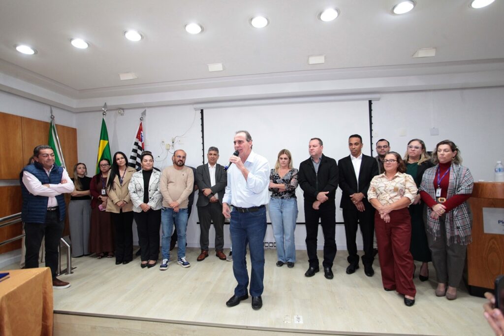 A imagem mostra um grupo de pessoas reunidas em um auditório ou sala de reuniões, provavelmente durante um evento oficial ou corporativo. * No centro da cena, um homem de camisa azul clara e calça jeans está de pé, segurando um microfone e falando para o público. * Ao seu redor, há cerca de 13 pessoas, homens e mulheres, dispostas em uma linha semicircular, observando o orador. Eles vestem trajes que variam do casual ao social. * O ambiente possui um piso de madeira clara e paredes brancas. Ao fundo, à esquerda, há mastros com a bandeira do Brasil e a bandeira do estado de São Paulo. * O teto é equipado com várias luminárias embutidas que iluminam o espaço de forma uniforme. À direita, parte de um púlpito de madeira é visível.