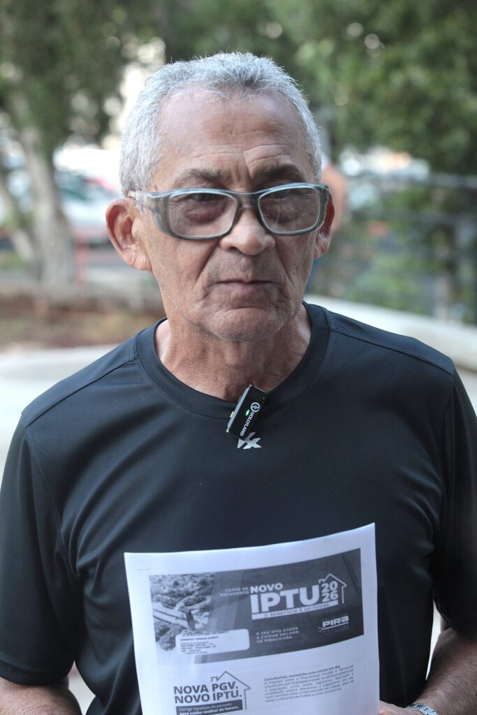 A imagem apresenta um homem de meia-idade, com cabelos curtos e grisalhos, de frente para a câmera. Ele usa óculos de grau com armação clara e uma camiseta preta de tecido esportivo. Preso à gola da camisa, há um pequeno microfone de lapela da marca Hollyland. O homem segura um documento impresso em papel branco, posicionado na altura do peito. O documento exibe o texto "NOVO IPTU 2026" e o logotipo da prefeitura de Piracicaba, indicando que se trata de uma comunicação oficial sobre o imposto predial. O fundo da imagem é desfocado, sugerindo um ambiente externo com vegetação e claridade natural.