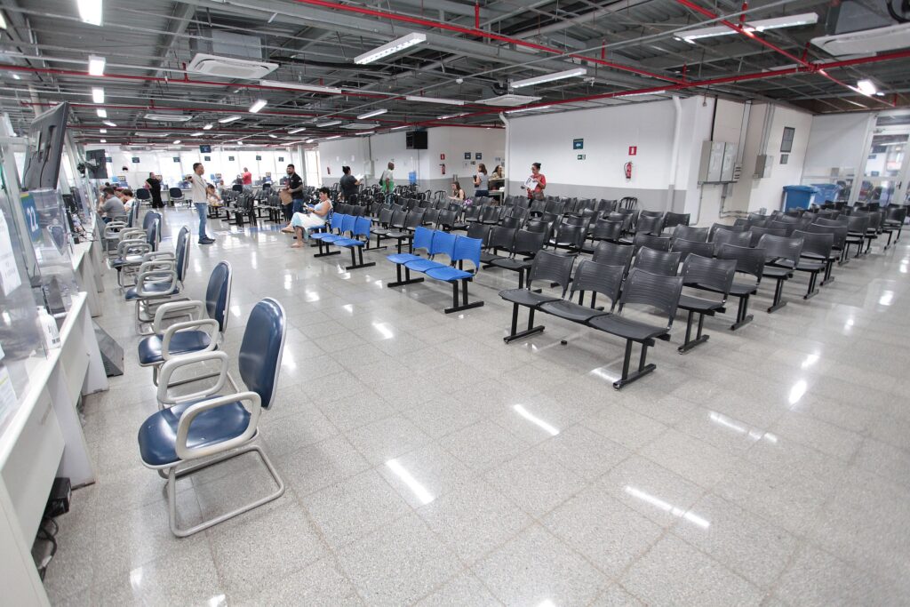Esta imagem mostra um amplo salão de espera, possivelmente de um órgão público ou repartição administrativa, com um design funcional e industrial. * O ambiente possui um teto alto com tubulações expostas, iluminação fluorescente linear e piso de granito polido que reflete as luzes. * À esquerda, há uma bancada de atendimento com guichês separados por divisórias de acrílico. Cadeiras individuais com braços estão alinhadas paralelamente a esta bancada. * O restante do salão é preenchido por várias fileiras de cadeiras de espera metálicas, a maioria na cor preta, com exceção de um conjunto de cadeiras azuis localizado mais à frente. * Algumas pessoas estão sentadas ou caminhando pelo espaço, indicando que o local está em funcionamento. O ambiente é bem iluminado, organizado e transmite uma atmosfera de repartição pública.