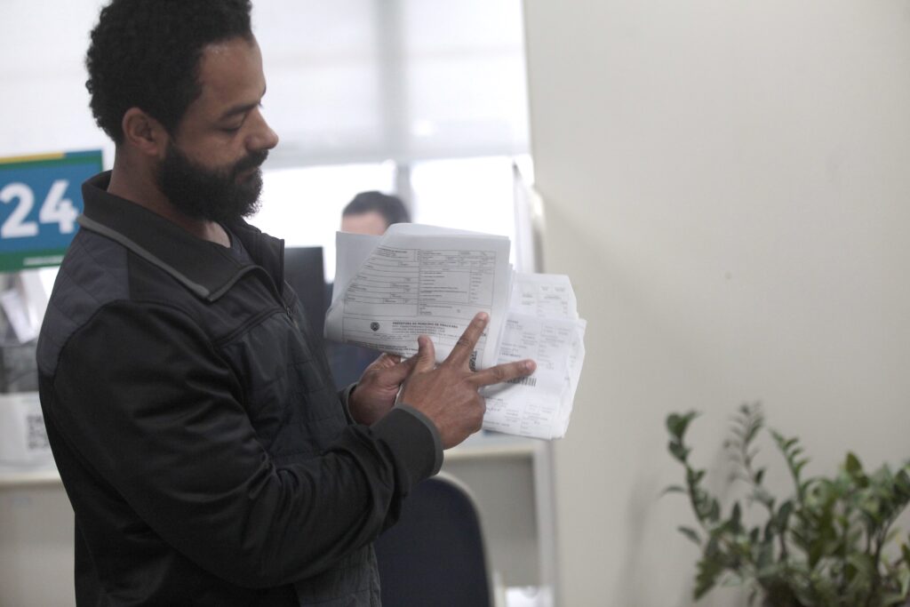 A imagem mostra um homem de pele parda, com barba e cabelo escuros, vestindo uma jaqueta preta. Ele está de perfil, voltado para a esquerda, segurando um conjunto de documentos ou boletos de cobrança com as duas mãos. O homem aponta com o dedo indicador da mão direita para um dos documentos, que apresenta o timbre da "Prefeitura do Município de Piracicaba". O ambiente parece ser um escritório ou repartição pública, com uma placa de sinalização contendo o número "24" visível ao fundo. À direita, há uma planta decorativa sobre uma superfície, e o foco da imagem está concentrado no homem e nos papéis que ele analisa.
