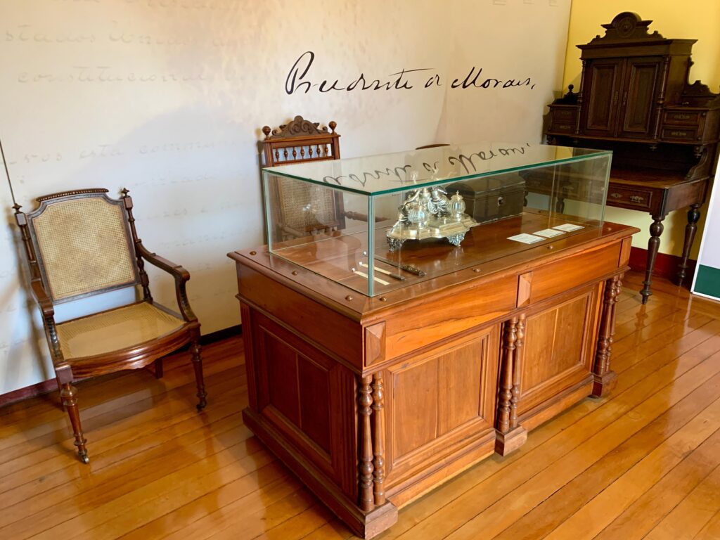 A imagem mostra um ambiente histórico, possivelmente uma sala de estudo ou escritório de época, que parece estar preservado em um museu. No centro, há uma grande mesa de madeira antiga com um tampo de vidro, onde estão expostos objetos, como um tinteiro prateado e uma pequena caixa escura. À esquerda da mesa, há uma cadeira antiga de madeira com encosto e assento de palhinha, típica do século XIX. Ao fundo, à direita, há um móvel alto e ornamentado, que parece ser um armário ou escrivaninha antiga, também de madeira escura e com detalhes clássicos. Na parede ao fundo, em letras cursivas grandes, está escrito "Prudente de Morais", que indica que o ambiente está relacionado a essa figura histórica, provavelmente o terceiro presidente do Brasil. O piso é de madeira polida, refletindo a luz suave que ilumina o espaço, conferindo um ar sóbrio e elegante ao ambiente. A composição geral sugere um local de trabalho ou estudo de uma personalidade importante, preservado para fins culturais e históricos.