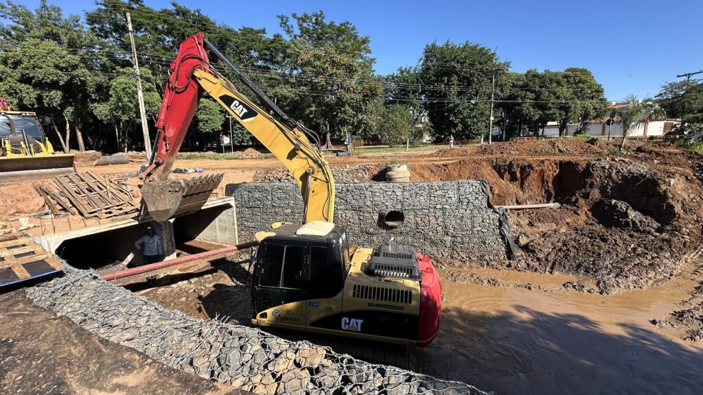 A imagem mostra um canteiro de obras ao ar livre, em um dia claro com céu azul. No centro da cena, há uma escavadeira grande da marca Caterpillar (CAT), com partes amarelas e vermelhas, posicionada em um terreno escavado e parcialmente alagado com água barrenta. A escavadeira está próxima a uma estrutura de concreto que parece ser um bueiro ou canal de drenagem. Ao lado direito da máquina, há uma parede de gabiões (estruturas de malha metálica preenchidas com pedras) que serve como contenção do terreno. Um trabalhador, usando capacete e camisa de manga longa, está dentro ou perto da abertura da estrutura de concreto, provavelmente inspecionando ou realizando algum serviço. Ao fundo, vê-se vegetação verde e postes de energia, indicando que o local está em uma área semi-rural ou próxima a uma zona urbana. A imagem transmite a ideia de uma obra de infraestrutura, possivelmente relacionada a drenagem ou contenção de solo.