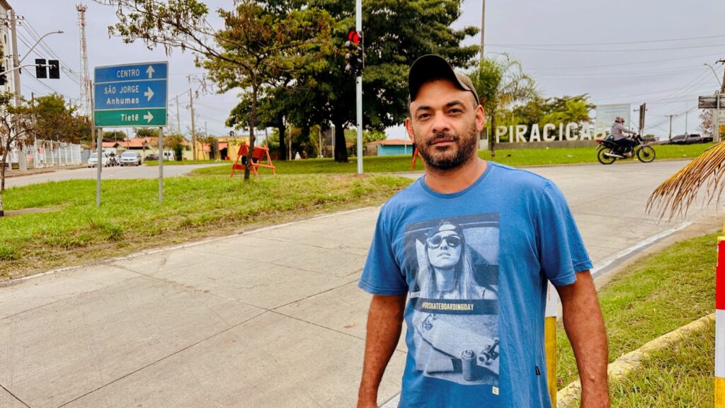 A imagem mostra um homem em um ambiente urbano ao ar livre, possivelmente em uma calçada próxima a uma rua ou cruzamento. Ele está de pé, olhando diretamente para a câmera, vestindo uma camiseta azul com uma estampa colorida que inclui a frase "#GOSKATEBOARDINGDAY" e a imagem de uma pessoa usando óculos escuros e boné. Ele também usa um boné escuro. Ao fundo, há uma placa grande com o nome "PIRACICABA" em letras maiúsculas, indicando que a foto foi tirada nessa cidade brasileira. Também é possível ver uma placa de sinalização verde com indicações para "CENTRO", "SÃO JORGE", "ANHUMAS" e "TIETÊ", sugerindo que o local é uma interseção que direciona para diferentes bairros ou cidades próximas. Há ainda um semáforo e a presença de algumas árvores e grama, indicando uma área com vegetação urbana. Um motociclista aparece ao fundo, mostrando que a via está em uso. O clima parece nublado, pois a luz está difusa, sem sombras fortes. A foto é tirada na altura dos olhos, capturando o homem da cintura para cima e proporcionando uma boa visão do cenário ao redor.