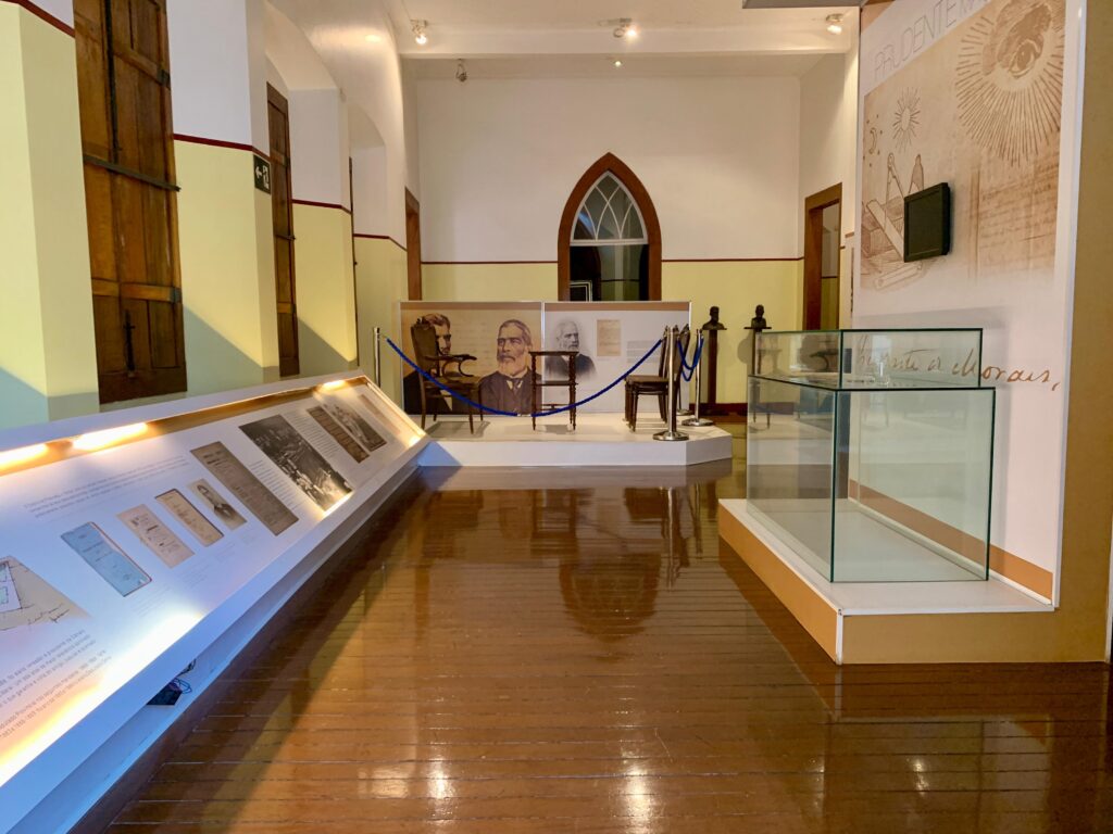A imagem mostra o interior de um museu ou espaço expositivo histórico, provavelmente no Brasil, dado o texto em português. O ambiente é organizado de forma simétrica, com um corredor central de chão de madeira polida que reflete a iluminação suave do teto. À esquerda, há uma grande vitrine inclinada contendo documentos históricos, fotografias e painéis explicativos. À direita, uma vitrine de vidro grande e vazia está posicionada sobre uma plataforma elevada. Ao fundo, no centro, um espaço protegido por cordões exibe uma cadeira de madeira antiga, uma pequena mesa, retratos de um homem com barba, e duas pequenas esculturas em pedestal. As paredes são pintadas em tons claros, divididas horizontalmente por uma faixa vermelha fina, e há uma janela arqueada ao fundo. Na parede direita, um painel gráfico grande exibe a palavra "PRUDENTE" acompanhada da frase "virtude e morais", além de desenhos que remetem a um olho e ferramentas geométricas, sugerindo um tema de sabedoria, ética e conhecimento. A iluminação é quente e indireta, criando um ambiente tranquilo e propício à contemplação histórica. A composição da imagem enfatiza a profundidade do espaço e a organização cuidadosa dos objetos expostos, transmitindo um ambiente cultural e educativo.