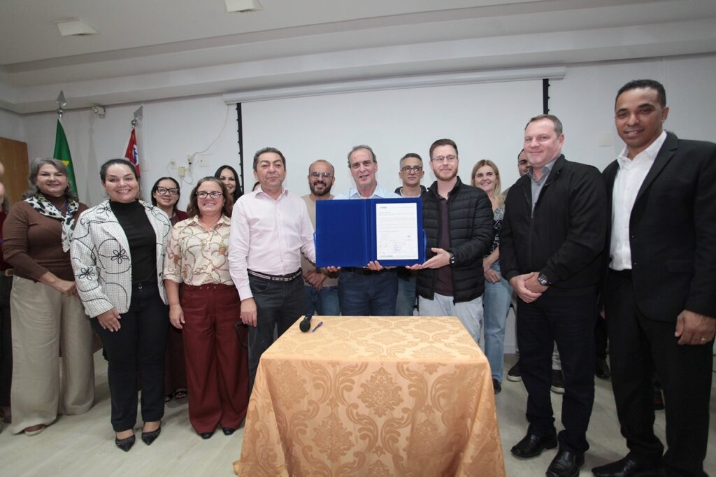 A imagem mostra um grupo de cerca de 13 pessoas, composto por homens e mulheres, posando para uma foto oficial em um ambiente interno, possivelmente uma sala de reuniões ou auditório. * No centro, dois homens seguram uma pasta azul aberta, que contém um documento oficial com assinaturas. * As pessoas estão vestidas de forma formal ou casual elegante. À esquerda, as mulheres usam blazers, calças sociais e camisas. Os homens no centro e à direita vestem camisas sociais, blazers ou jaqueta. * À frente do grupo, há uma mesa coberta por uma toalha de tecido bege com detalhes em relevo, onde repousa uma caneta. * O ambiente é iluminado, com paredes brancas e uma tela de projeção enrolada ao fundo. À esquerda, é possível ver parte de duas bandeiras, sendo uma delas a bandeira do Brasil. O clima parece ser de uma cerimônia de assinatura de convênio, posse ou algum evento institucional.