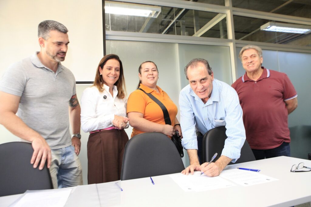 A imagem mostra um grupo de cinco pessoas em um ambiente interno, provavelmente um escritório ou sala de reunião. No primeiro plano, um homem está assinando um documento sobre uma mesa branca. Ele veste uma camisa azul clara xadrez e parece estar sorrindo. Ao seu lado, uma mulher com camiseta polo laranja e uma bolsa preta está observando a assinatura. Perto dela, outra mulher, vestindo uma camisa branca e saia marrom, também sorri enquanto olha para o homem que está assinando. À esquerda da imagem, um homem com camiseta cinza e bermuda jeans está em pé, com o braço apoiado em uma cadeira preta, olhando para a mesa. Ao fundo, há outro homem com camiseta polo vermelha, que também observa a cena e sorri. O ambiente é iluminado por luzes fluorescentes no teto, e ao fundo há divisórias de vidro fosco, sugerindo um espaço corporativo. A atmosfera parece ser de formalidade e satisfação, possivelmente durante a assinatura de um contrato ou acordo.