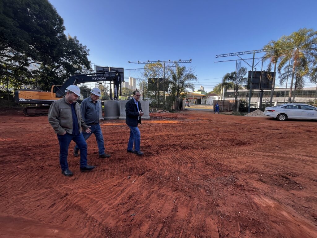 A imagem mostra um canteiro de obras ao ar livre em um dia ensolarado com céu azul claro. No centro, há uma grande área de terra vermelha, revolvida e com marcas de pneus. Três homens caminham sobre essa área: dois deles usam capacetes brancos e roupas de trabalho, enquanto o terceiro está vestido de forma mais casual, com jaqueta e calça jeans. À esquerda, há uma escavadeira hidráulica da marca Hyundai, modelo 220LC-9, estacionada próxima a algumas barreiras de concreto. Ao fundo, é possível ver uma cerca de arame farpado, algumas árvores, um posto de gasolina e um carro branco estacionado. A cena sugere uma inspeção ou avaliação do andamento da obra.