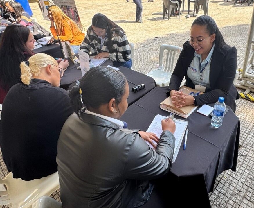 A imagem mostra um evento ao ar livre, possivelmente uma feira de empregos ou uma sessão de orientação, com várias mesas organizadas. No foco principal, duas mulheres estão interagindo em uma mesa coberta por um tecido preto. A mulher à direita, que usa óculos e veste um blazer escuro sobre uma camisa azul clara com um logo, está sorrindo e parece estar conduzindo uma entrevista ou ajudando a outra mulher. A mulher à esquerda, vista de costas, está vestindo uma jaqueta preta e está escrevendo em um documento apoiado em uma prancheta. Sobre a mesa, há papéis, canetas, uma garrafa de água e um pequeno bloco de notas. Ao fundo, outras mesas e pessoas indicam que o evento é maior. A iluminação é natural, sugerindo que é dia, e o ambiente parece ser um espaço aberto com piso pavimentado e algumas estruturas que podem ser tendas ou estandes. A cena transmite uma atmosfera profissional e organizada, com um momento de interação formal entre as participantes.A imagem mostra um evento ao ar livre, possivelmente uma feira de empregos ou uma sessão de orientação, com várias mesas organizadas. No foco principal, duas mulheres estão interagindo em uma mesa coberta por um tecido preto. A mulher à direita, que usa óculos e veste um blazer escuro sobre uma camisa azul clara com um logo, está sorrindo e parece estar conduzindo uma entrevista ou ajudando a outra mulher. A mulher à esquerda, vista de costas, está vestindo uma jaqueta preta e está escrevendo em um documento apoiado em uma prancheta. Sobre a mesa, há papéis, canetas, uma garrafa de água e um pequeno bloco de notas. Ao fundo, outras mesas e pessoas indicam que o evento é maior. A iluminação é natural, sugerindo que é dia, e o ambiente parece ser um espaço aberto com piso pavimentado e algumas estruturas que podem ser tendas ou estandes. A cena transmite uma atmosfera profissional e organizada, com um momento de interação formal entre as participantes.