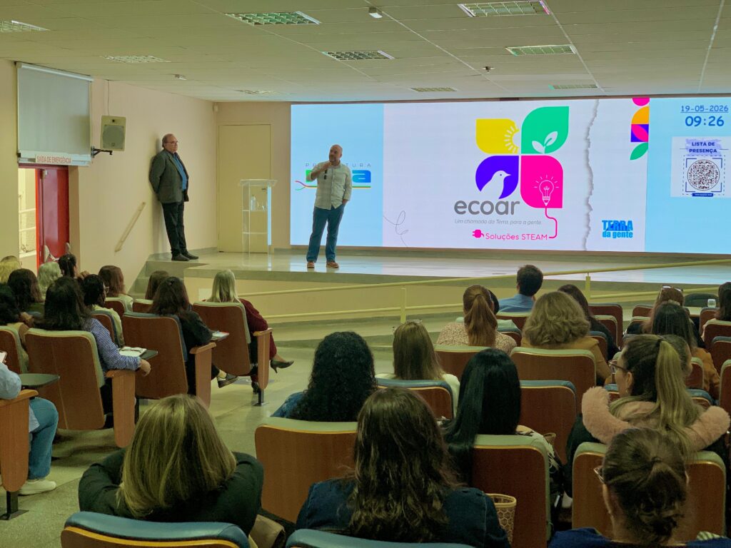 A imagem mostra uma palestra ou apresentação acontecendo em um auditório moderno. No palco, há dois homens: um está falando para a plateia, enquanto o outro está de pé próximo à parede, observando. Atrás deles, um grande telão exibe o logo colorido da iniciativa "ecoar", com o slogan "Um chamado da Terra, para a gente", e a frase "Soluções STEAM". Também aparece a data "19-05-2026", o horário "09:26" e um QR code para a lista de presença. A plateia está sentada em cadeiras estofadas dispostas em fileiras, assistindo atentamente à apresentação. O ambiente é iluminado por luzes de teto, com paredes claras, transmitindo um clima profissional e educacional. Na parte superior do telão, há a indicação de uma saída de emergência e a palavra "Prefeitura", sugerindo que o evento pode estar relacionado a uma iniciativa pública ou municipal.