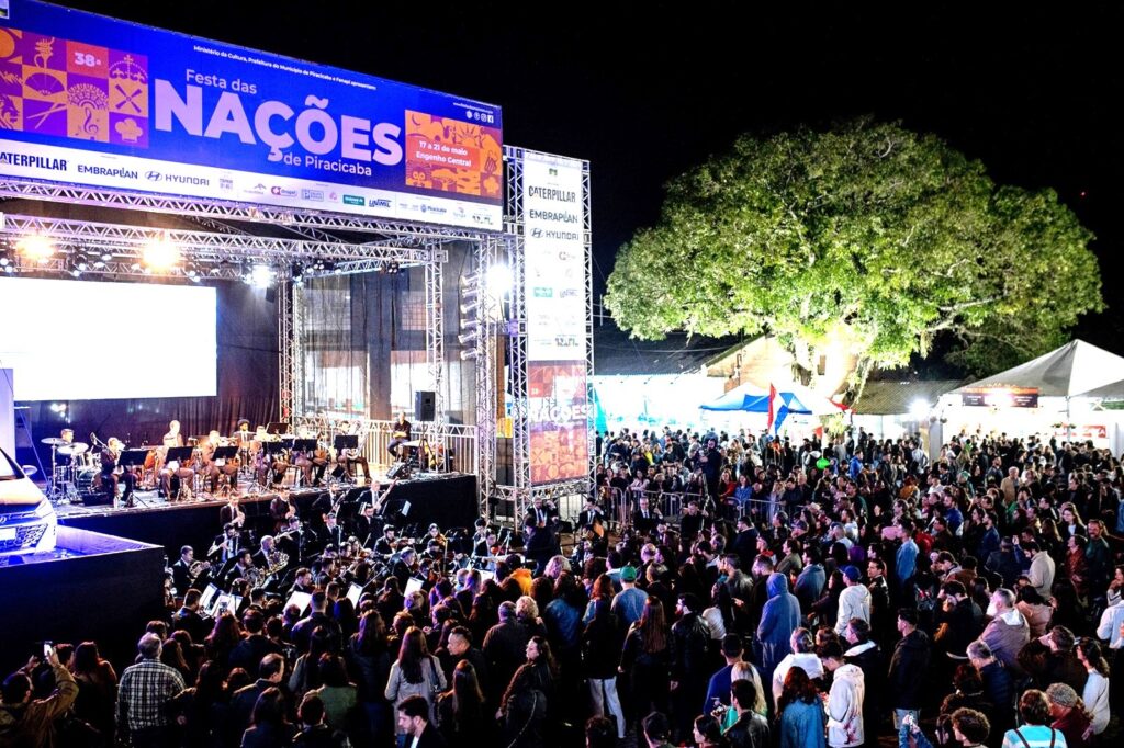 A imagem mostra um evento ao ar livre realizado à noite, chamado "38ª Festa das Nações de Piracicaba". No centro da imagem, há um grande palco elevado onde uma orquestra está se apresentando. O palco é estruturado com uma armação metálica e tem uma grande faixa azul e laranja na parte superior com o nome do evento e os logos de patrocinadores como Caterpillar, Embraplan e Hyundai. Na frente do palco, uma grande multidão está reunida, assistindo ao espetáculo. As pessoas estão bastante próximas umas das outras, indicando um público expressivo. À direita da imagem, há uma árvore iluminada e algumas tendas brancas, sugerindo que o evento oferece outras atrações além da música. O cenário é noturno, com o palco bem iluminado, destacando os músicos, enquanto o público está em uma área mais escura. A atmosfera parece festiva e cultural, com muita gente participando e aproveitando o evento. A imagem foi tirada de uma posição elevada, proporcionando uma visão ampla do palco e da plateia.