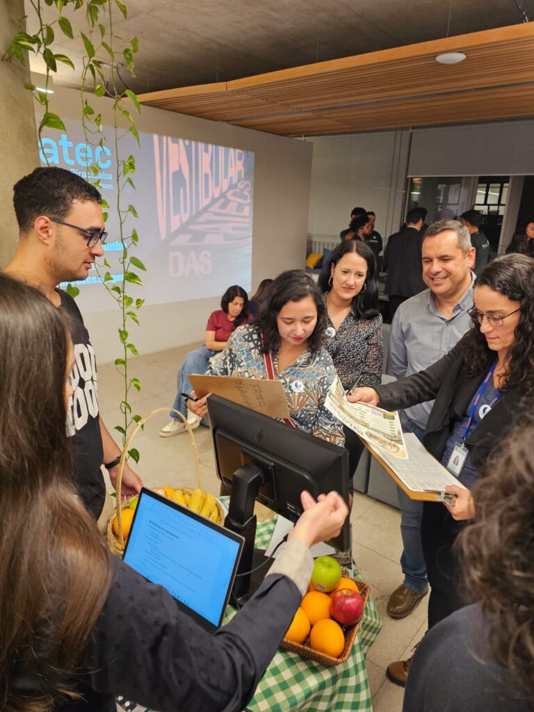 Esta imagem registra um momento em um ambiente que parece ser uma feira de profissões, evento acadêmico ou apresentação de projeto da FATEC (Faculdade de Tecnologia), realizado em Piracicaba. Ambiente: Espaço moderno de convivência com teto de concreto aparente e ripas de madeira. Ao fundo há projeção na parede com os dizeres "VESTIBULAR FATEC" e "DAS". Uma planta pendurada (jibóia) decora uma coluna à esquerda. Pessoas: Grupo de aproximadamente sete a oito pessoas em torno de uma mesa. À esquerda, um jovem de óculos e camiseta preta parece explicar algo. No centro, uma mulher com camisa estampada segura uma placa de papelão onde se lê "FATEC PIRACICABA". À direita, uma mulher de óculos e blazer preto segura um documento ou panfleto, consultando-o com um homem de camisa cinza e jeans. Outras pessoas aparecem ao fundo e em primeiro plano. Mesa e objetos: Mesa com toalha xadrez verde e branca. Sobre a mesa, dois cestos de vime com frutas (bananas, laranjas e maçãs), sugerindo tema ligado à nutrição, agricultura, tecnologia de alimentos ou sustentabilidade. Tecnologia: Monitor de computador sobre a mesa voltado ao grupo e um notebook aberto em primeiro plano sendo operado por uma pessoa; a tela mostra um documento de texto ou planilha. Atmosfera: A imagem transmite trabalho em equipe, ambiente acadêmico-profissional e uma apresentação ou avaliação de projeto.