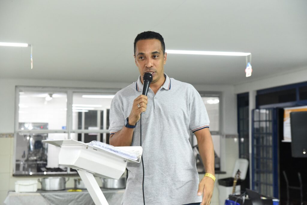 A imagem mostra um homem falando ao microfone, aparentemente fazendo uma apresentação ou discurso. Ele está vestido com uma camisa polo cinza clara com detalhes nas cores vermelho, branco e azul na gola. O homem tem cabelo curto e escuro, pele clara e usa um relógio preto no pulso esquerdo, além de um anel prateado no dedo. Ele está atrás de um púlpito branco e parece estar em um ambiente interno, possivelmente uma cozinha industrial ou refeitório, dado que ao fundo há panelas grandes e utensílios de cozinha. A iluminação é clara e o ambiente tem janelas grandes, com paredes brancas e estrutura metálica aparente no teto. A expressão do homem é séria e concentrada, indicando que ele está focado no que está dizendo.