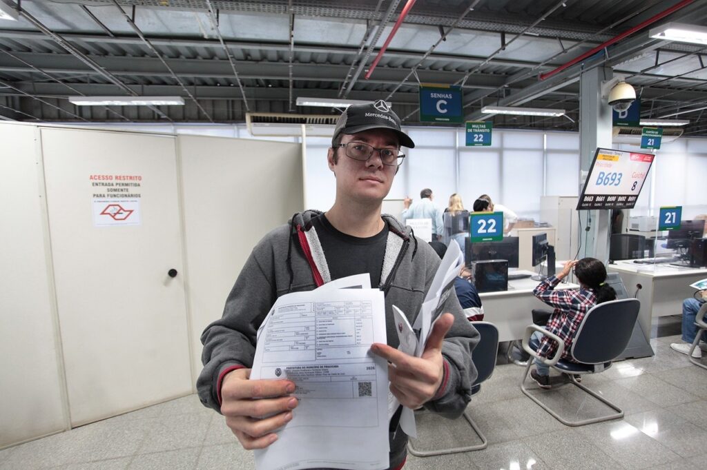A imagem mostra um jovem em primeiro plano, posicionado em um ambiente que parece ser um centro de atendimento público ou órgão governamental, possivelmente em Piracicaba, dado o documento que segura. * O homem veste um moletom cinza, óculos de grau e um boné da marca Mercedes-Benz. Ele segura papéis, que parecem ser guias de pagamento de impostos municipais (como IPTU), e aponta para um deles com a mão esquerda. * Ao fundo, o local é um escritório de atendimento com divisórias e guichês. Há placas indicativas no teto, como "SENHAS C", "MULTAS DE TRÂNSITO" e números de guichês (21 e 22). * À direita, uma tela exibe o número de senha "B693" sendo atendido no guichê 8. Outras pessoas estão sentadas ou em pé, aguardando atendimento. * À esquerda, há uma porta branca com um aviso de "ACESSO RESTRITO" e um logotipo da prefeitura local. O ambiente é iluminado por luzes fluorescentes de teto, conferindo um aspecto funcional e administrativo ao local.