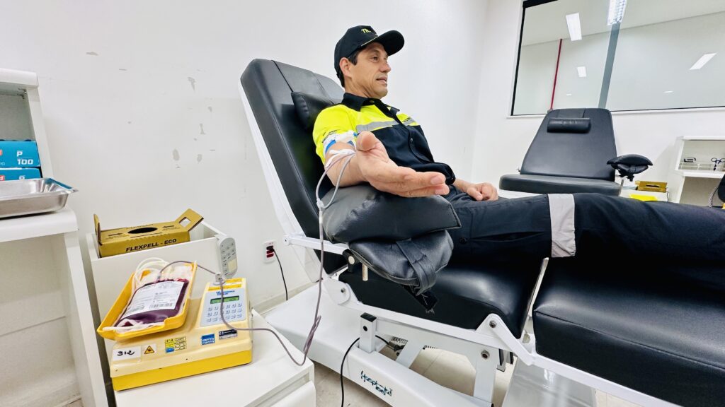 A imagem mostra um homem em um centro de coleta de sangue, sentado em uma poltrona hospitalar reclinável, realizando uma doação de sangue. * O homem, que veste um uniforme de trabalho e um boné, está com o braço direito estendido, onde está inserido o acesso venoso conectado a um tubo. * Ao lado da poltrona, sobre uma mesa branca, há um equipamento médico amarelo (uma balança homogeneizadora de sangue) que está conectado à bolsa de coleta. A bolsa, contendo sangue, repousa sobre a bandeja do equipamento. * O ambiente é clínico, com paredes brancas, uma segunda poltrona vazia ao fundo e suprimentos médicos, como luvas, visíveis em uma prateleira lateral. A cena transmite um momento de solidariedade e cuidado com a saúde.