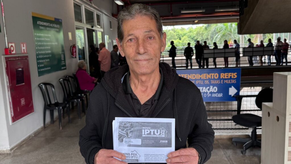 A imagem apresenta um homem de meia-idade, com cabelos grisalhos e vestindo um casaco preto com capuz, posicionado no centro em primeiro plano. Ele segura um documento impresso com o título "CARNÊ DE PAGAMENTO NOVO IPTU 2026", referente à Prefeitura de Piracicaba. O cenário é um ambiente de atendimento público, possivelmente um centro administrativo municipal. Ao fundo, à esquerda, há uma placa na parede listando serviços como "Atendimento Tributário" e "Multas de Trânsito". À direita, observa-se uma placa azul com os dizeres "ATENDIMENTO REFIS" e uma indicação para o "9º ANDAR". O ambiente é iluminado por luz natural e artificial, com diversas pessoas sentadas ou em pé ao fundo, aguardando atendimento. O foco principal é o homem, que olha diretamente para a câmera, enquanto o restante da cena compõe o contexto de um órgão público.