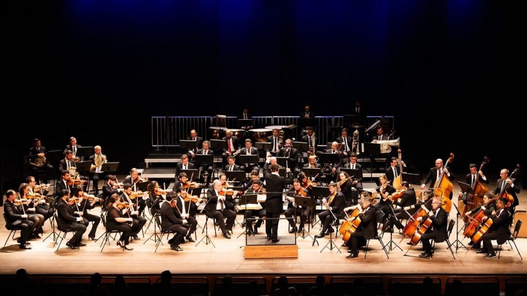 A imagem mostra uma orquestra sinfônica em pleno concerto, vista de um ângulo amplo que abrange todo o palco. No centro, um maestro está de pé sobre um pequeno estrado, conduzindo os músicos. À frente e no meio do palco, estão os instrumentos de corda, como violinos, violas, violoncelos e contrabaixos, enquanto na parte de trás, em níveis mais elevados, estão os instrumentos de percussão e metais. Todos os músicos vestem roupas formais pretas, típicas de apresentações clássicas. O chão do palco é de madeira clara e polida, refletindo a luz que vem de cima, que ilumina os músicos e seus instrumentos, destacando o brilho das superfícies metálicas e das madeiras. O fundo é completamente preto, o que cria um contraste forte, focando toda a atenção nos artistas. Na parte inferior da imagem, é possível ver as silhuetas das cabeças do público, sugerindo que a foto foi tirada da plateia, dando uma sensação de imersão na apresentação. A atmosfera é formal, elegante e profissional, típica de um concerto clássico.