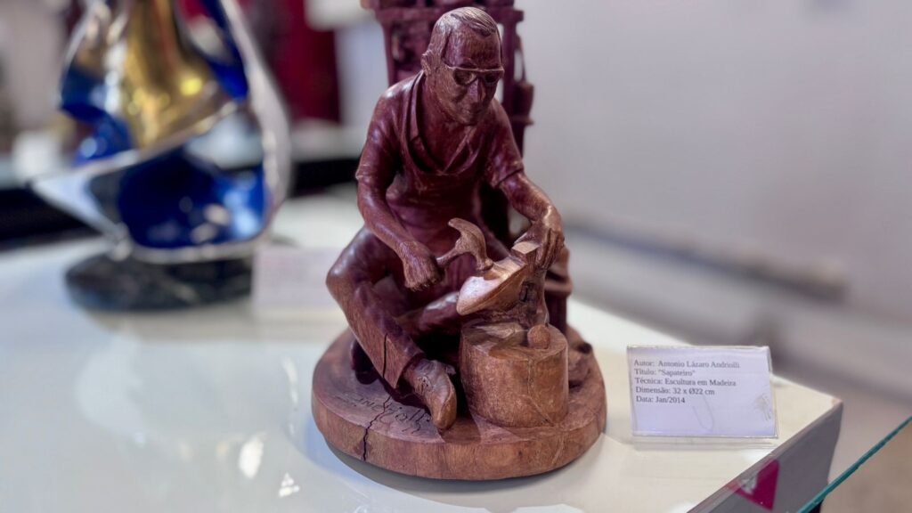 A imagem mostra uma escultura detalhada em madeira representando um sapateiro em atividade. A figura é de um homem mais velho, usando óculos e avental, sentado em um banquinho baixo. Ele está concentrado trabalhando em um sapato, segurando uma ferramenta, possivelmente um martelo, sobre uma forma de sapato apoiada em um bloco de madeira. A escultura é rica em detalhes, evidenciando as dobras da roupa, os traços do rosto e a textura da madeira, que tem um tom avermelhado. Ao lado da escultura, há uma plaquinha branca com informações sobre a obra, indicando que o autor é Antonio Lázaro Andriolli, o título é "Sapateiro", a técnica utilizada é escultura em madeira, as dimensões são 32 cm de altura por 22 cm de diâmetro, e a data da criação é janeiro de 2014. Ao fundo, desfocado, há outra escultura de estilo mais abstrato e metálico, de cor azul. A iluminação é suave, destacando bem os detalhes da escultura principal.