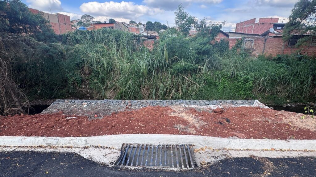 A imagem mostra uma paisagem com um pequeno riacho que atravessa uma área semiurbana ou residencial. De um lado do riacho, há uma vegetação densa composta por capim alto e outras plantas verdes. Do outro lado, há uma parede de gabião (estrutura feita com pedras dentro de uma malha de arame) que parece ter sido construída para proteger a margem contra erosão. Ao lado dessa parede, há uma área de terra exposta, com solo de coloração avermelhada, indicando que pode estar em processo de construção ou recuperação. Ao fundo, é possível ver casas de tijolos com telhados vermelhos, típicas de um bairro residencial simples. Também há uma estrada asfaltada à direita da imagem, onde um carro branco está estacionado ou passando. A iluminação é natural, com céu claro, sugerindo que a foto foi tirada durante o dia. A cena transmite a ideia de uma localidade em desenvolvimento, com intervenções para controle ambiental e infraestrutura.