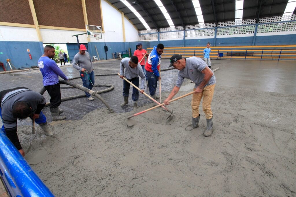 A imagem mostra uma equipe de construção realizando a concretagem do piso de uma quadra poliesportiva coberta. O grupo trabalha de forma coordenada para espalhar e nivelar o concreto fresco sobre uma malha de ferro, usando ferramentas de cabo longo. É um ambiente de ginásio, com estrutura de telhado visível, tabela de basquete ao fundo e arquibancadas laterais nas cores azul e amarela. Posso ajudar com mais alguma coisa em relação a esta obra, como detalhar os passos desse processo de concretagem ou elaborar um relatório sobre a cena?