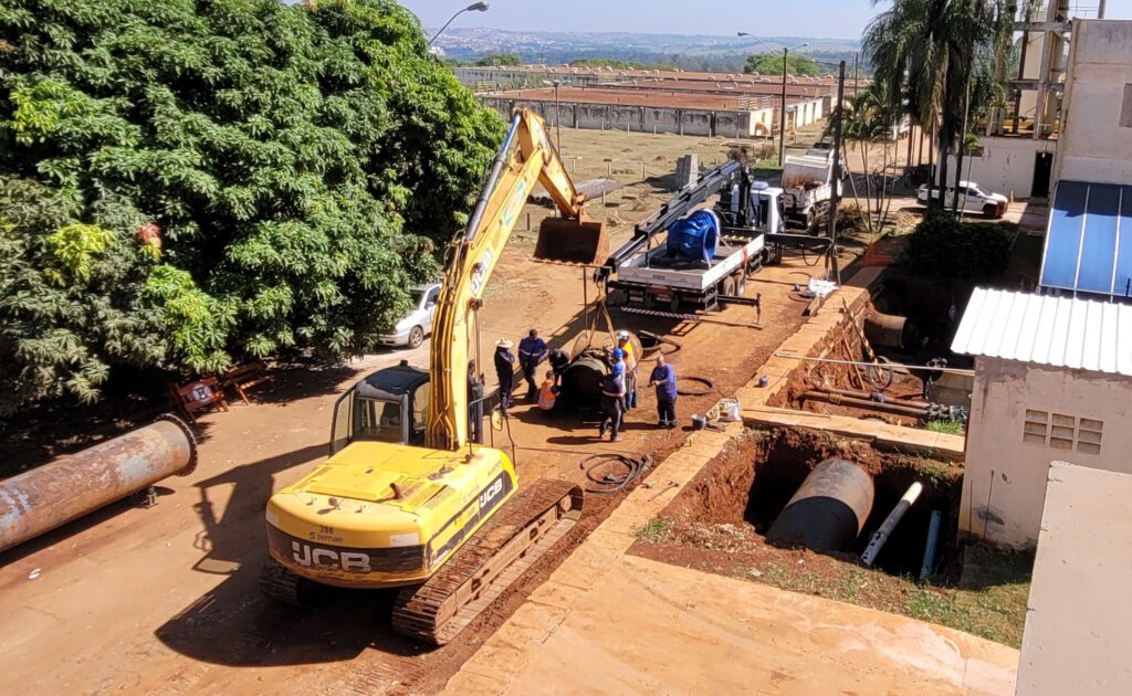 A imagem mostra um canteiro de obras ao ar livre visto de um ângulo elevado. No centro, um grupo de trabalhadores usando capacetes e equipamentos de segurança está reunido ao redor de uma grande tubulação cilíndrica escura. À esquerda, há uma escavadeira amarela da marca JCB, com o braço levantado. Perto dos trabalhadores, há um caminhão com um guindaste, que está carregando um componente industrial azul. O terreno é de terra, e à frente há uma escavação profunda onde outra seção de tubulação está visível. À esquerda da imagem, há uma árvore grande e verde, enquanto à direita há uma estrutura branca com um telhado azul que parece ser composto por painéis solares. Ao fundo, é possível ver edifícios industriais e uma paisagem distante sob um céu claro e ensolarado. A cena sugere uma atividade de construção ou manutenção de infraestrutura, com o uso de máquinas pesadas e trabalho em equipe.