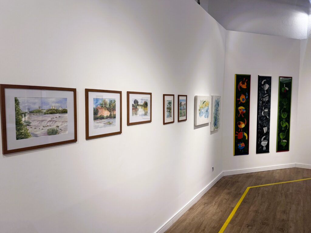A imagem mostra o interior de uma galeria de arte com paredes brancas e piso de madeira clara. Há uma linha amarela marcada no chão que segue uma curva ao longo da parede. Na parede à esquerda, estão penduradas várias obras de arte pequenas, principalmente pinturas emolduradas que parecem retratar paisagens. Mais à frente, na parede que faz uma curva, há três painéis verticais grandes e estreitos com arte abstrata: um verde com padrões circulares, outro escuro com formas circulares brancas, e um terceiro colorido com formas abstratas variadas. A iluminação é clara e uniforme, típica de um espaço de exposição. A perspectiva da foto é ligeiramente baixa, mostrando parte do chão e das obras na parede. No centro da imagem, está escrito em letras grandes e pretas: "VOCE TEM CASA PROPRIA".