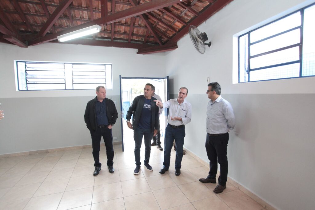 A imagem mostra quatro homens em um ambiente interno simples, possivelmente um escritório ou uma sala comunitária. As paredes são brancas com uma faixa cinza na parte inferior, o teto tem estrutura de madeira exposta e o chão é revestido com azulejos claros. Há duas janelas com moldura metálica, uma à esquerda e outra à direita, além de uma porta no centro que dá para o exterior. Os homens estão em pé, próximos uns dos outros, e parecem estar em uma conversa ou discussão. Um deles, o terceiro da esquerda para a direita, está gesticulando com a mão direita, como se estivesse explicando algo. Outro homem veste uma camiseta azul escura com inscrições, incluindo "Avan" e "JOÃO A.". A iluminação do ambiente é natural, entrando pelas janelas e pela porta aberta, complementada por uma luminária fluorescente no teto. Na parede à direita, há um ventilador de parede. O ambiente parece funcional e simples, com foco na interação dos homens.