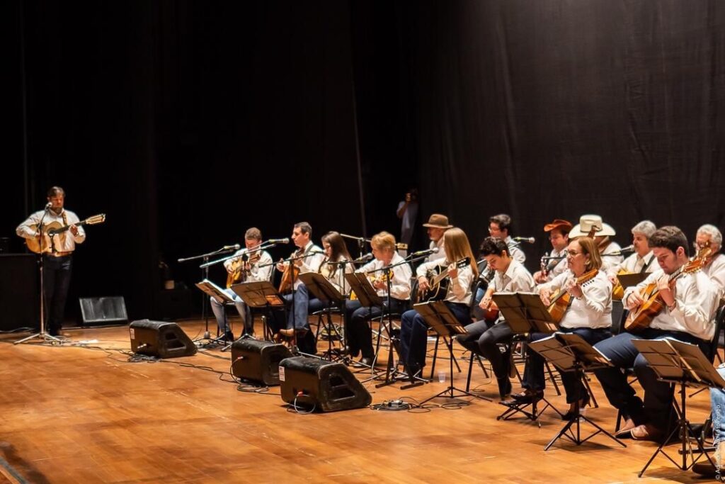 A imagem mostra um conjunto musical em um palco, composto por cerca de 15 músicos, a maioria tocando instrumentos de corda, como violões e possivelmente bandolins. Um músico está em pé à esquerda, parecendo liderar ou fazer um solo, enquanto os demais estão sentados em filas, cada um com um suporte para partituras à frente. Todos vestem camisas claras, predominantemente brancas, dando um aspecto uniforme e formal ao grupo. O palco está iluminado por luzes frontais que destacam os músicos e seus instrumentos, com um fundo escuro que ajuda a focar a atenção no conjunto. Na parte frontal do palco, há caixas de som pretas, indicando que se trata de uma apresentação ao vivo. A perspectiva da foto é do ponto de vista da plateia, capturando a amplitude do grupo e a atmosfera de um concerto formal.