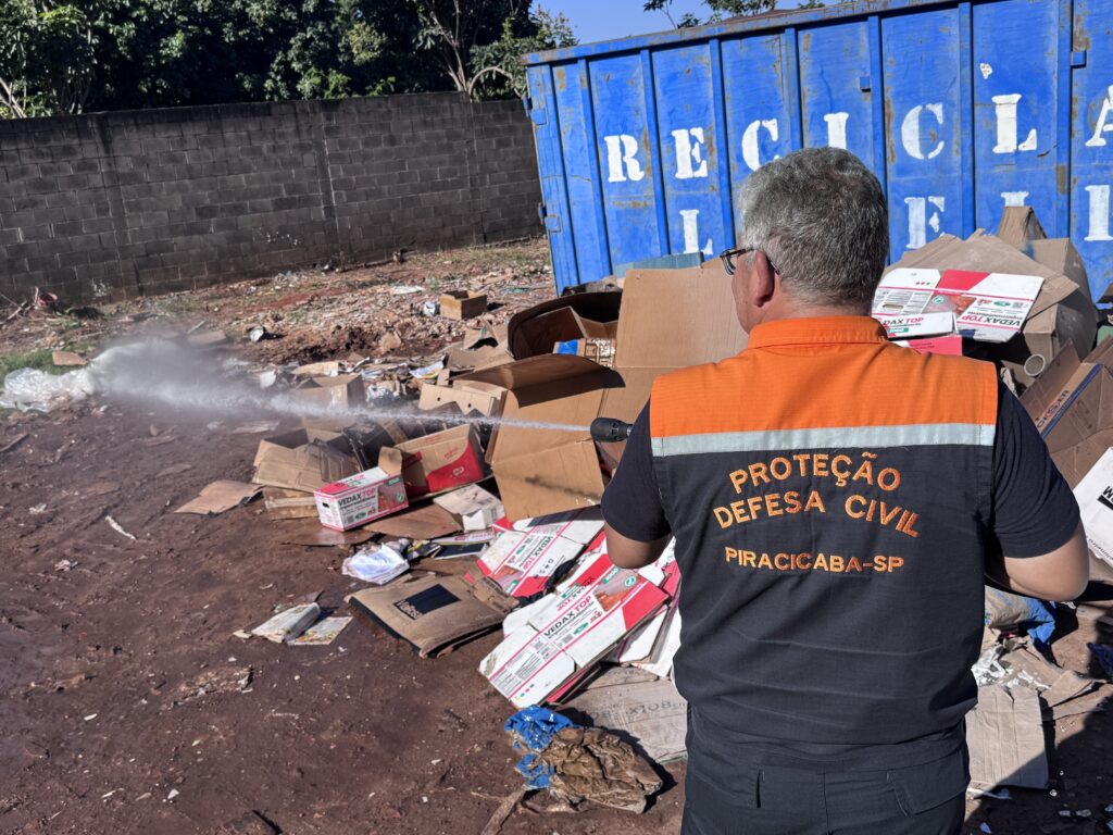 A imagem mostra um agente da Defesa Civil de Piracicaba-SP, vestindo um colete laranja e preto, de costas, utilizando um equipamento de pulverização para aplicar um produto sobre um amontoado de resíduos, principalmente caixas de papelão. * O agente está posicionado à direita, direcionando o jato de água ou produto químico para o centro de uma área aberta onde há diversos papéis e embalagens descartados. * Ao fundo, observa-se uma grande caçamba de metal azul com a inscrição "RECICLAGEM" parcialmente visível, além de um muro de blocos de concreto e vegetação sob um céu limpo e ensolarado. * O ambiente parece ser um local de descarte ou triagem de materiais recicláveis, onde a ação do agente sugere uma medida de controle, possivelmente relacionada à sanitização, combate a pragas ou prevenção de focos de incêndio em materiais inflamáveis.