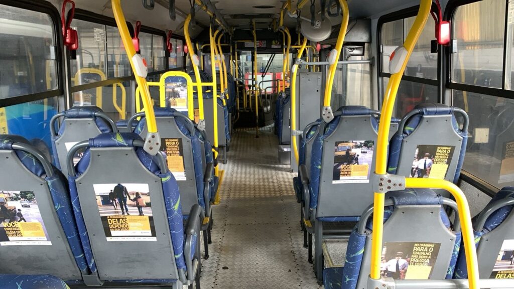A imagem mostra o interior de um ônibus público, visto de dentro, olhando para a frente. O corredor central está livre, com assentos azuis estampados alinhados dos dois lados, cada um com encosto de plástico cinza. Há barras e suportes amarelos para os passageiros se segurarem, distribuídos ao longo do corredor. O piso é metálico com um padrão texturizado, típico de veículos de transporte público. Nas costas dos assentos, há cartazes informativos em português, que trazem mensagens de conscientização sobre segurança no trânsito, especialmente para pedestres. Os textos principais dos cartazes dizem: "NO CAMINHO PARA O TRABALHO, NÃO DEIXE A PRESSA TE CEGAR" e "VOCÊ ENXERGA ALÉM DO CELULAR? A SEGURANÇA DELAS DEPENDE DA SUA VISÃO", alertando para os perigos de usar o celular e a importância de estar atento no trânsito. A iluminação do ônibus é clara, provavelmente natural, entrando pelas janelas, e o ambiente parece limpo e organizado. A foto é simétrica, com o foco centralizado no corredor que leva até a parte da frente do ônibus, onde o motorista está localizado.