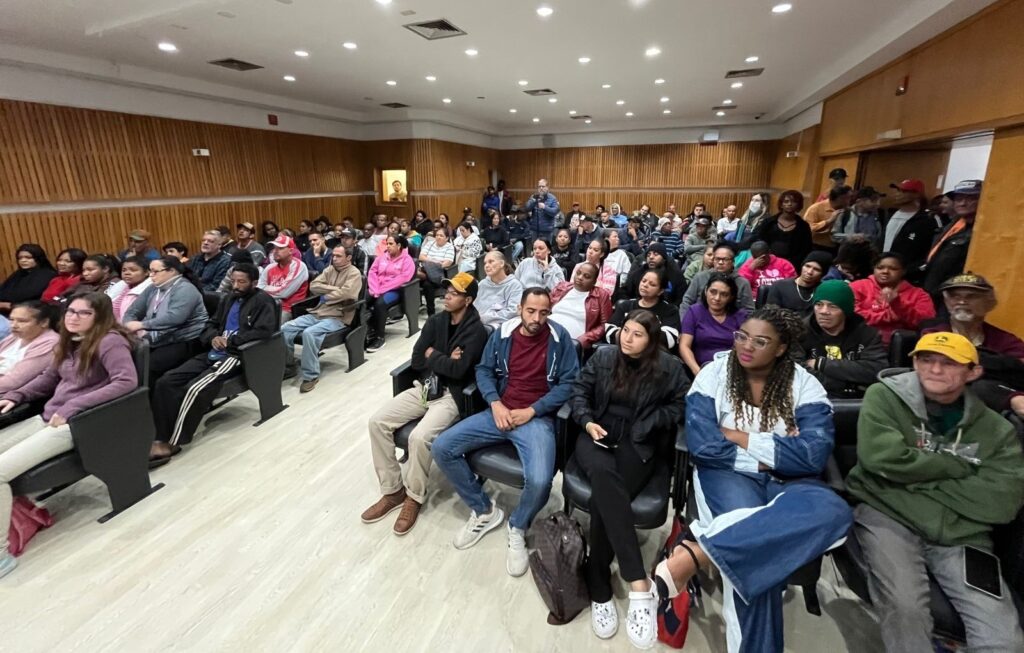 A imagem mostra uma grande sala de auditório ou um anfiteatro, repleto de pessoas sentadas em cadeiras organizadas em fileiras. A maioria das pessoas parece jovem e está vestida de forma casual, com roupas como moletons, jaquetas e camisetas. O ambiente é bem iluminado, com luzes embutidas no teto, que é branco e liso. As paredes laterais são revestidas com painéis de madeira clara, conferindo um aspecto moderno e acolhedor ao espaço. No fundo da sala, algumas pessoas estão em pé, possivelmente organizadores ou palestrantes. A expressão das pessoas é majoritariamente atenta, sugerindo que estão assistindo a uma palestra, apresentação ou evento educacional. A atmosfera geral é de concentração e interesse.