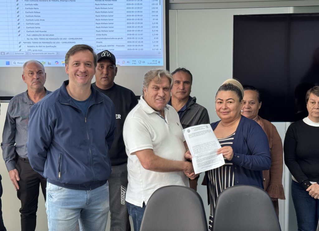 A imagem mostra um grupo de oito pessoas em um ambiente de escritório, provavelmente relacionado à Prefeitura do Município de Piracicaba, conforme indicado pelo documento que uma mulher segura. No centro da imagem, um homem de camisa polo branca está cumprimentando uma mulher que veste blusa listrada e um cardigã escuro, que segura um papel com o brasão da prefeitura. Ao fundo, há outras seis pessoas, algumas sorrindo e outras com expressões mais neutras, todas olhando para a câmera. Na parede atrás deles, uma grande tela exibe uma lista de arquivos ou e-mails relacionados a currículos, liberação de recursos e outros documentos administrativos. O ambiente é iluminado e formal, sugerindo um momento de entrega ou apresentação oficial de documentos.