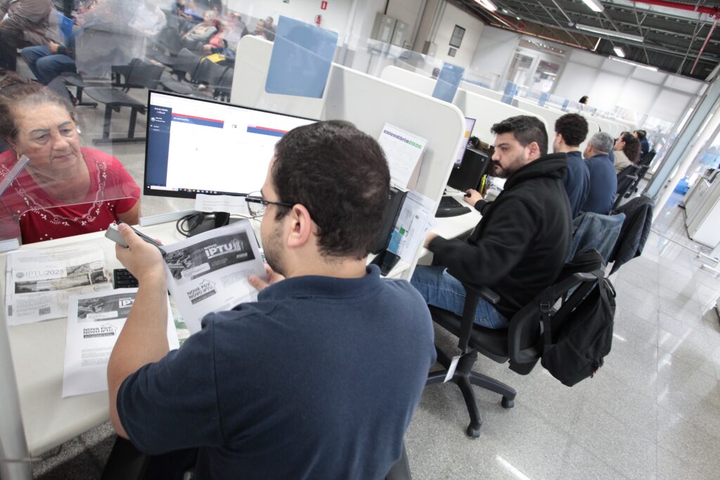 A imagem mostra um ambiente de atendimento ao público, possivelmente uma repartição pública ou setor de impostos. * Em primeiro plano, um atendente jovem, vestindo uma camisa polo azul escura, está sentado à mesa, manuseando papéis que contêm a inscrição IPTU. Ele está de costas para a câmera, voltado para uma mulher idosa que está do outro lado de uma divisória de acrílico transparente. * A mulher, que veste uma blusa vermelha, observa atentamente o atendente enquanto ele organiza os documentos. À frente do atendente, há um monitor de computador e outros documentos impressos sobre a mesa. * Ao fundo, em uma disposição de escritório em linha, outros atendentes estão sentados em suas estações de trabalho, focados em seus computadores. O ambiente possui um piso de granito claro e divisórias brancas entre as mesas, criando um espaço de trabalho organizado e profissional.