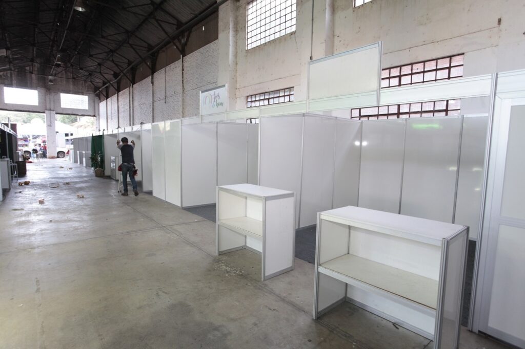 A imagem mostra o interior de um grande galpão industrial, possivelmente um centro de exposições ou feira, em processo de montagem para um evento. No primeiro plano, há dois balcões modulares brancos, feitos com estrutura de alumínio e painéis brancos, que estão vazios. Ao fundo, uma longa fileira de estandes também modulares está sendo montada, todos brancos e alinhados lado a lado. Um trabalhador está em um pequeno escada, aparentemente ajustando ou instalando algum componente dos estandes. O ambiente tem um piso de concreto e o teto alto, com estrutura metálica aparente e grandes janelas com grades, que deixam entrar bastante luz natural. No painel de um dos estandes está escrito "Fios do Bem". Ao fundo, é possível ver a entrada ou saída do galpão, com iluminação natural vinda do lado de fora. No geral, a imagem transmite a sensação de preparação e organização para um evento em um espaço amplo e industrial.