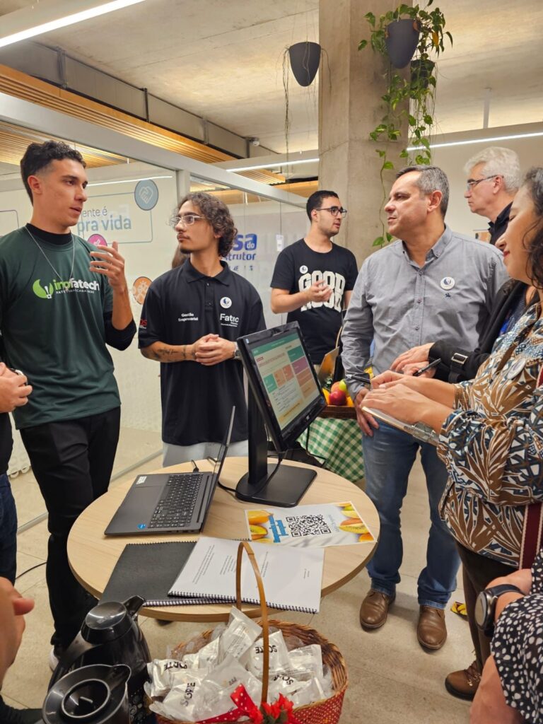 A imagem mostra um ambiente interno, possivelmente uma feira, evento acadêmico ou profissional, onde um grupo de pessoas está reunido em torno de uma mesa redonda. À esquerda, um jovem veste uma camiseta verde com a palavra "Infatech" e parece estar explicando algo com gestos. Ao lado dele, outro jovem com cabelo encaracolado e óculos, usando uma camisa preta com o logo "Fatec", escuta atentamente. Do outro lado da mesa, um homem mais velho, vestido com camisa cinza e calça jeans, segura uma caneta e um bloco de anotações, demonstrando interesse na conversa. Ao seu lado, uma mulher com blusa estampada também segura um caderno, provavelmente anotando informações. Na mesa, há um laptop aberto, um monitor exibindo algum tipo de software, um folheto com um código QR e uma cesta com embalagens, possivelmente de brindes ou snacks. O ambiente é moderno, com paredes de vidro, piso claro e luminárias no teto. Também há uma coluna de concreto com um vaso de planta pendurado, conferindo um toque de verde ao espaço. Ao fundo, mais pessoas estão presentes, e um jovem com camiseta preta com a palavra "GOOD" está visível. No geral, a cena transmite uma atmosfera de interação profissional ou acadêmica, com troca de informações e demonstração de produtos ou projetos.