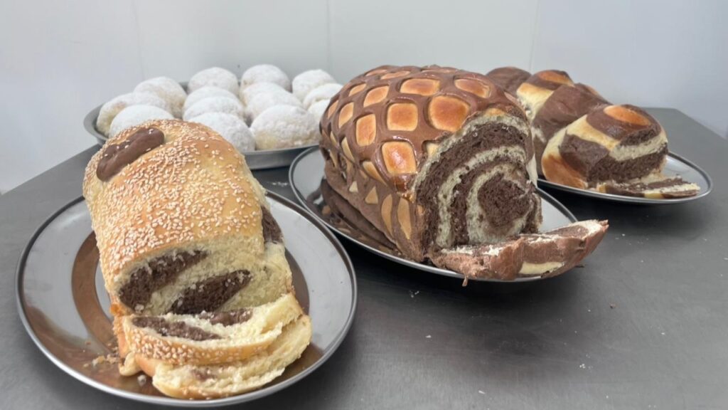 A imagem mostra uma variedade de pães e doces fresquinhos dispostos sobre uma superfície metálica, provavelmente em uma padaria ou cozinha. Em destaque, há três tipos principais de pães: À esquerda, um pão com sementes de gergelim por cima, com um recheio visível de chocolate, cortado para mostrar o interior. No centro, um pão com uma crosta trabalhada em um padrão geométrico, lembrando uma treliça, também com recheio de chocolate, parcialmente fatiado. À direita, um pão trançado ou enrolado, mesclando massa clara e recheio de chocolate. Ao fundo, há uma bandeja redonda com vários pãezinhos pequenos cobertos por açúcar de confeiteiro, parecendo macios e doces. A luz é clara e realça as texturas crocantes da casca dos pães e o aspecto macio e açucarado dos pãezinhos ao fundo. A perspectiva é levemente aérea, dando uma boa visão geral dos produtos. A composição é harmoniosa, com os itens bem distribuídos na imagem.