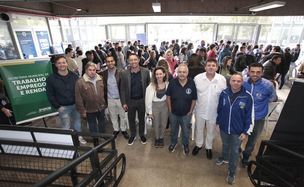 A imagem mostra um grupo de pessoas posando para uma foto em um evento público, que parece ser uma ação de serviços comunitários, possivelmente uma feira de empregos ou atendimento municipal em Piracicaba, Brasil. Aqui estão os detalhes principais: O Grupo: As oito pessoas aparecem organizadas em uma linha, vestindo roupas casuais a formais, e parecem ser os organizadores ou autoridades presentes no evento. Contexto: Ao fundo, vemos um espaço amplo com bastante movimento, onde muitas outras pessoas circulam, o que reforça a ideia de um evento com público. Local: É possível identificar uma placa verde da "Secretaria Municipal de Trabalho, Emprego e Renda" e um banner do "Procon", indicando que o local oferece serviços voltados ao trabalhador e ao consumidor. A estrutura parece ser um galpão ou espaço industrial com teto alto e bastante luminosidade natural. A cena transmite um tom de colaboração e organização. Parece ser o registro de um momento importante durante a realização de um serviço público para a comunidade.