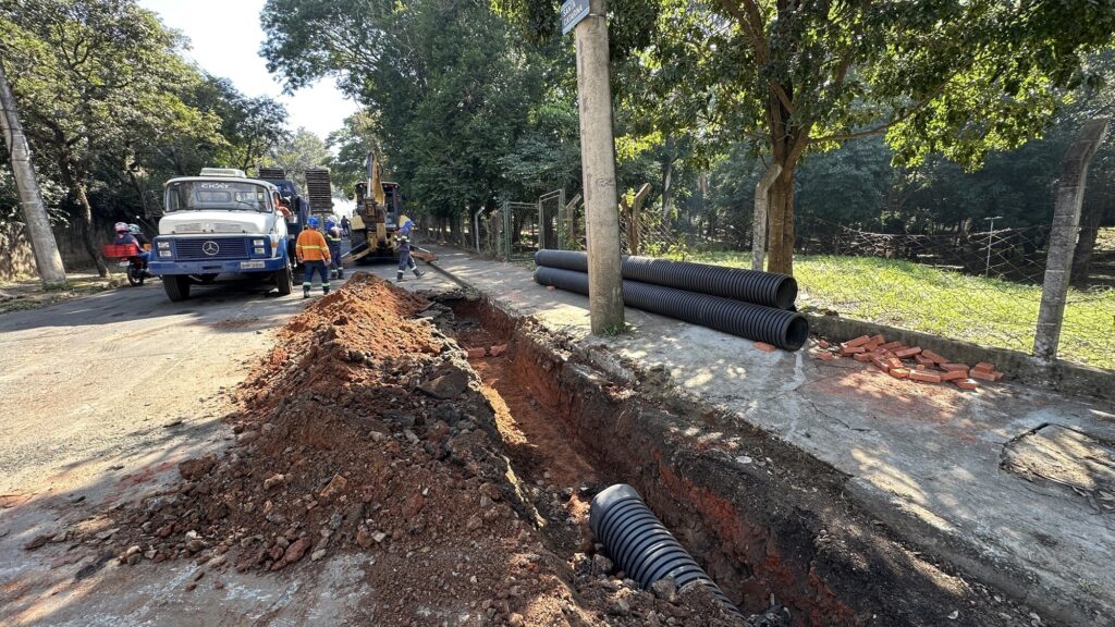 A imagem mostra uma obra em andamento ao ar livre, em um dia ensolarado. Um trecho da calçada e da rua está aberto, com uma vala escavada onde tubos de drenagem pretos, corrugados, estão sendo instalados. Há uma grande quantidade de terra removida ao lado da vala. Um caminhão azul da empresa "CICAT" está estacionado próximo à obra, e à frente dele há uma escavadeira amarela. Três operários, todos usando coletes refletivos, capacetes e roupas de proteção, estão trabalhando no local. Ao fundo, há árvores verdes e uma cerca de arame. Um poste de concreto com uma placa indicando "SANTA CATARINA" está em evidência à direita da imagem. O céu está claro e azul, indicando um dia ensolarado. A cena transmite uma sensação de atividade e trabalho em infraestrutura urbana.