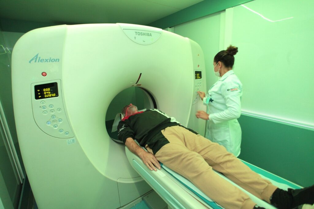 A imagem mostra uma cena clínica dentro de uma sala de exames médicos, provavelmente para uma tomografia computadorizada (TC). O foco principal é um aparelho grande e branco da marca Toshiba, modelo Alexion, que ocupa a maior parte do lado esquerdo e central da imagem. Um paciente do sexo masculino está deitado de costas na mesa motorizada do aparelho, com a cabeça posicionada dentro do anel do scanner. Ele veste uma camiseta preta com algumas inscrições brancas e calça de moletom bege. À direita do aparelho, uma técnica em saúde, usando jaleco branco, calça escura e máscara facial branca, está de pé, operando o painel de controle do equipamento. Seu cabelo está preso em um coque. A sala é bem iluminada, com uma luz fria que realça o ambiente clínico e esterilizado. As paredes são verdes claras. No equipamento, há displays digitais mostrando números, como 00.0, 046 e 0284.0, provavelmente relacionados aos parâmetros do exame. A imagem é uma tomada ampla, capturada de um ângulo levemente elevado, mostrando claramente a interação entre o paciente, a técnica e o equipamento médico.