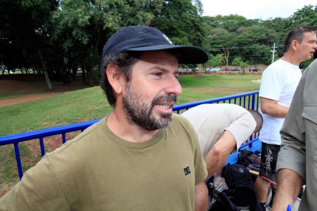 A imagem mostra uma cena ao ar livre, provavelmente em um parque ou área verde. Em primeiro plano, há um homem de meia-idade, com barba e cabelo escuro, usando um boné preto e uma camiseta verde oliva com o logo "DC" no peito. Ele está olhando para o lado direito da imagem, com uma expressão tranquila e um leve sorriso. Ao seu lado direito, parcialmente visível, há outro homem vestindo camiseta branca e shorts pretos, também olhando na mesma direção. Mais à direita, aparece parte de uma terceira pessoa com camiseta bege. Ao fundo, vê-se uma cerca azul de metal, uma área gramada com algumas árvores e uma rua com carros estacionados ou em movimento. A iluminação é natural, indicando um dia nublado ou com luz suave. A cena transmite um ambiente descontraído e casual, possivelmente durante um passeio ou encontro informal.