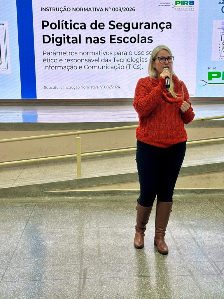 A imagem mostra uma mulher em pé, falando ao microfone, provavelmente em um evento ou apresentação. Ela veste um suéter laranja de tricô grosso, calça escura e botas marrons até o joelho. Ela usa óculos e tem cabelo loiro. Ao fundo, há um telão com um slide projetado que traz o título "INSTRUÇÃO NORMATIVA N° 003/2026" e o tema "Política de Segurança Digital nas Escolas", que trata de normas para o uso seguro, ético e responsável das Tecnologias de Informação e Comunicação (TICs). O slide também informa que esta instrução substitui a anterior, número 002/2024. No canto superior direito do telão, está o logo da Secretaria de Educação de Piracicaba, indicando que o evento é promovido por essa instituição. O ambiente parece ser uma sala ou auditório institucional, com piso brilhante e um corrimão ao fundo.