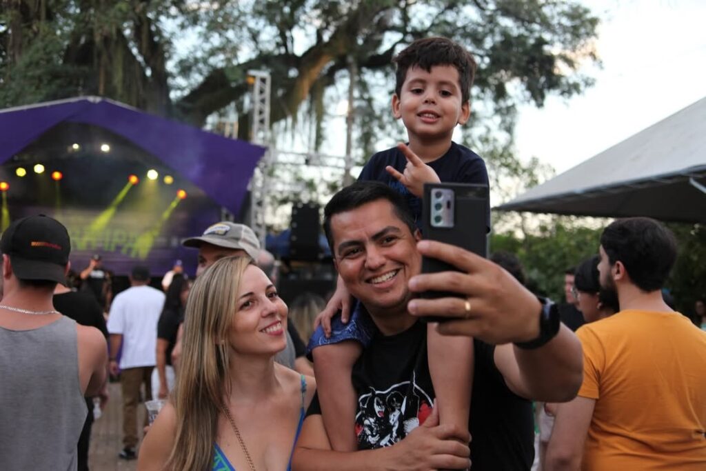 A imagem captura um momento de descontração em um evento ao ar livre, possivelmente um show ou festival. * Em primeiro plano, um homem sorridente segura um smartphone para tirar uma selfie, enquanto carrega uma criança nos ombros. A criança, também sorridente, faz um gesto com as mãos. * Ao lado do homem, uma mulher loira olha para a câmera com um sorriso suave. * O cenário ao fundo mostra um palco com iluminação cênica, estruturas metálicas e uma multidão de pessoas circulando, indicando um ambiente de festa. * A iluminação sugere o final da tarde ou início da noite, com um céu aberto e árvores ao fundo, compondo um ambiente festivo e familiar.
