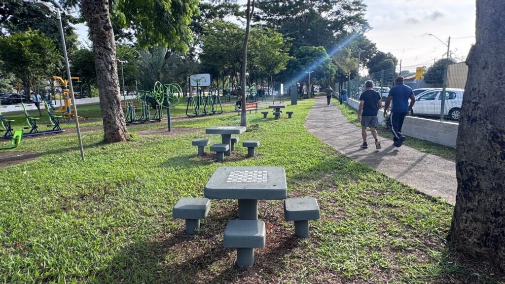 A imagem mostra um parque ao ar livre em um dia ensolarado. Em primeiro plano, há duas mesas de concreto com tabuleiros de jogo, possivelmente para damas ou xadrez, acompanhadas de pequenos bancos fixos ao redor. O gramado está bem cuidado e verde. Mais ao fundo, há uma área de exercícios com aparelhos de ginástica ao ar livre, como bicicletas ergométricas e outros equipamentos para atividades físicas. Um caminho pavimentado corta a cena, por onde duas pessoas caminham, uma vestindo camiseta escura e shorts e a outra, uma camisa polo azul com calça escura. No fundo, é possível ver árvores e uma área cercada com carros estacionados. O ambiente transmite uma sensação de tranquilidade e lazer, típico de um parque comunitário em um dia claro e ensolarado.