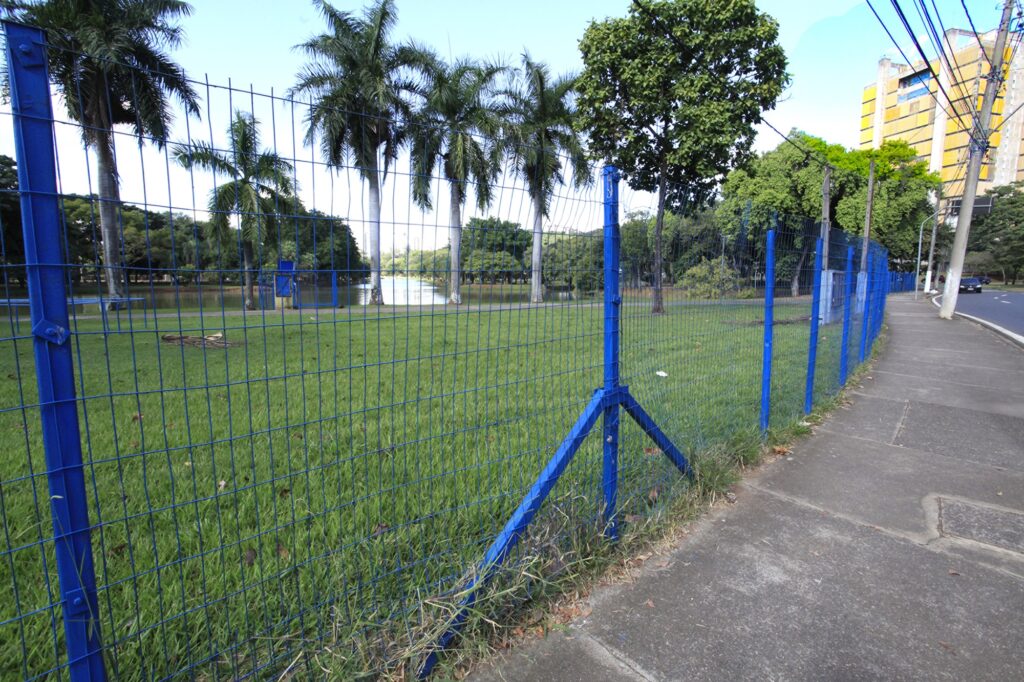 A imagem mostra uma cena ao ar livre, provavelmente um parque público, vista a partir de uma calçada ao lado de uma cerca azul de metal com malha. No centro da imagem há uma área verde com grama bem cuidada e várias árvores altas, incluindo palmeiras e outras árvores com folhagem densa. Ao fundo, há um pequeno corpo de água que reflete o céu claro e azul. A calçada de concreto segue paralela à cerca azul, que se destaca na imagem com sua cor vibrante e estrutura diagonal. À direita, ao fundo, é possível ver um prédio alto de cor amarela, sugerindo uma área urbana próxima. O céu está claro e ensolarado, o que indica um dia de clima agradável. A composição da imagem cria um contraste interessante entre o ambiente natural do parque e a estrutura urbana da calçada e do prédio.