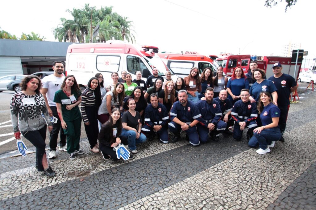 A imagem mostra um grande grupo de pessoas reunidas ao ar livre, posando para uma foto em um ambiente que parece ser uma área pavimentada, possivelmente uma rua ou estacionamento com piso de pedra. O grupo é composto por indivíduos usando uniformes do SAMU (Serviço de Atendimento Móvel de Urgência) e também por outras pessoas vestidas com roupas casuais, algumas com maquiagem que simula ferimentos, indicando que provavelmente participaram de um treinamento ou simulação de atendimento emergencial. Ao fundo, é possível ver veículos de emergência, incluindo ambulâncias do SAMU e um caminhão dos Bombeiros, ambos com os números "192" visíveis, que é o número de emergência no Brasil. A luz natural indica que a foto foi tirada durante o dia. Algumas pessoas seguram placas ou sinais azuis em forma de mão, sugerindo que fazem parte de uma campanha ou atividade de conscientização. No geral, a imagem transmite um ambiente de colaboração entre profissionais de emergência e a comunidade, possivelmente durante um exercício de primeiros socorros, resgate ou treinamento de atendimento a vítimas.
