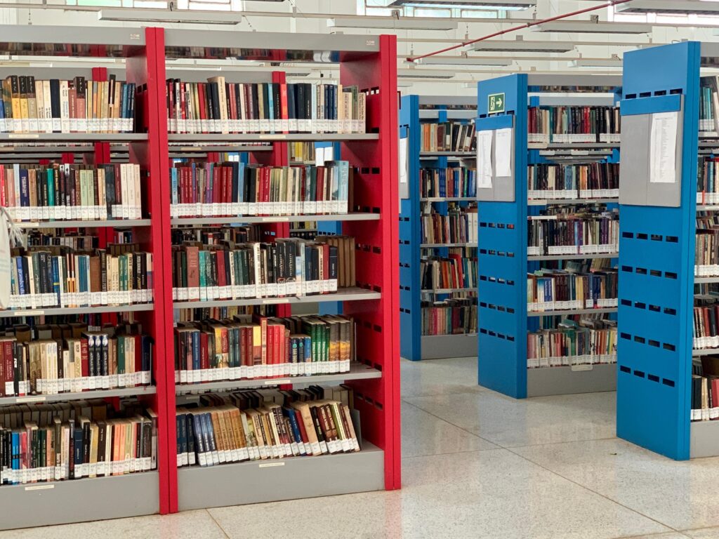 A imagem apresenta uma vista interna de uma biblioteca organizada, focada em várias estantes de livros dispostas em fileiras sobre um piso de granito polido. * Em primeiro plano, à esquerda, destaca-se uma estante com estruturas laterais na cor vermelha, densamente preenchida com livros de diversas cores e tamanhos em várias prateleiras. * Ao fundo e à direita, observa-se outra fileira de estantes, estas com estruturas laterais na cor azul, que seguem o mesmo padrão de organização. Nas laterais dessas estantes azuis, há placas brancas afixadas, possivelmente contendo informações ou listas de classificação. * O ambiente é iluminado por luzes artificiais instaladas no teto, que possui uma estrutura industrial aparente. A perspectiva da imagem cria um efeito de profundidade, guiando o olhar através dos corredores formados pelas estantes, transmitindo uma atmosfera de organização e quietude típica de um ambiente acadêmico ou de leitura.