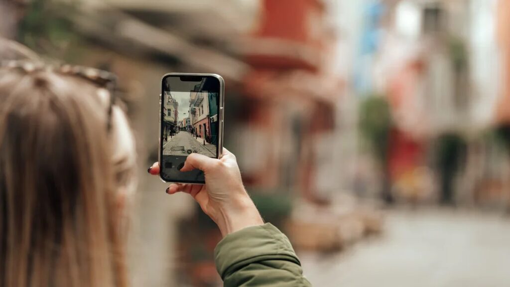 A imagem mostra uma pessoa vista por trás, segurando um smartphone para fotografar uma rua estreita e histórica. * A pessoa, com cabelos loiros, está em primeiro plano, com o braço estendido segurando o aparelho. * Na tela do celular, é possível ver a imagem capturada: uma rua de paralelepípedos ladeada por edifícios coloridos, típicos de vilas europeias. * O fundo da cena real está com um efeito de desfoque (bokeh), mantendo o foco total no celular e na mão da pessoa, destacando a ação de registrar o momento.