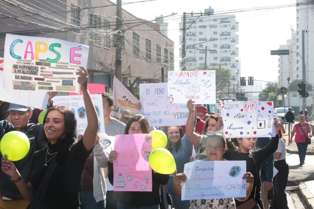 A imagem retrata uma manifestação pública ao ar livre, durante o dia, focada na causa da saúde mental e na luta antimanicomial. Um grupo de diversas pessoas caminha pela rua, muitas delas segurando cartazes feitos à mão e balões amarelos. Os participantes exibem mensagens de apoio aos CAPS (Centros de Atenção Psicossocial). Alguns dos dizeres nos cartazes incluem: * "CAPS É...": acompanhado de palavras como "potencial", "realidade", "se abrir", "inovar", "energia" e "crescer". * "Só o amor liberta": com desenhos e frases como "mente livre" e "viva o CAPS". * "Quem ama não tranca": enfatizando uma abordagem de cuidado sem isolamento. * "Luta antimanicomial - Trancar não é tratar": acompanhado de um desenho de uma gaiola aberta. * "Empatia + Preconceito - Respeito - Julgamento": destacando valores positivos em oposição aos negativos. O clima parece ser de engajamento e conscientização, com os manifestantes sorridentes e unidos em prol de um modelo de cuidado humanizado e em liberdade. Ao fundo, percebe-se o ambiente urbano de uma cidade, com prédios, fiação elétrica e semáforos.