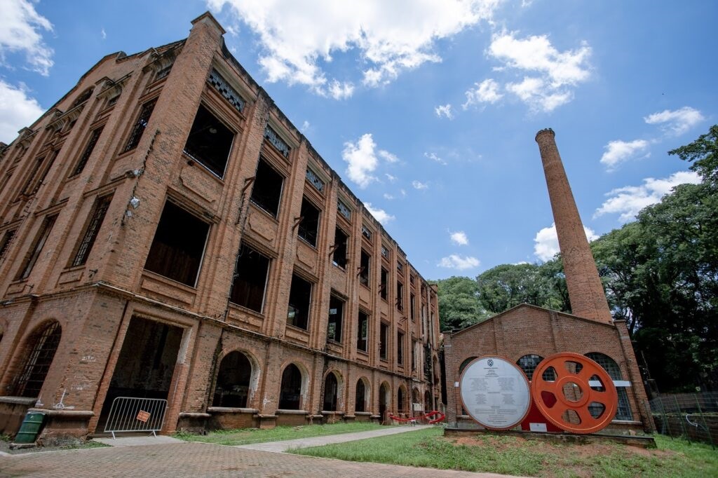 A imagem exibe as ruínas imponentes de uma antiga fábrica de tijolos aparentes, caracterizada por uma arquitetura industrial clássica. O edifício principal possui múltiplos andares com grandes aberturas de janelas vazias, sugerindo um estado de abandono ou preservação histórica. * Ao lado da estrutura principal, destaca-se uma chaminé alta e cilíndrica, típica de complexos industriais do século passado. * No primeiro plano, à direita, há dois elementos circulares em destaque: um painel informativo circular e uma grande engrenagem metálica pintada de laranja, que servem como monumentos ou referências ao passado fabril do local. * O cenário é composto por um gramado bem cuidado, um caminho pavimentado e um céu azul com nuvens dispersas, criando um contraste entre a estrutura histórica envelhecida e o ambiente externo preservado.