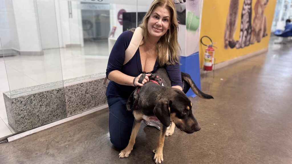 A imagem mostra uma mulher de cabelos loiros, sorrindo e ajoelhada no chão, enquanto abraça um cachorro de porte médio. O cão tem pelagem curta, predominantemente preta com manchas castanhas nas patas e no rosto, e está usando um peitoral vermelho. A mulher veste uma blusa azul-marinho de decote em V e calças escuras, carregando uma mochila bege nas costas. Eles estão em um ambiente interno que parece ser um corredor de um estabelecimento comercial, possivelmente um pet shop ou clínica veterinária, dado o fundo com paredes amarelas decoradas com ilustrações de animais e a presença de um extintor de incêndio. O piso é de concreto polido e há uma divisória de vidro ao lado da mulher.